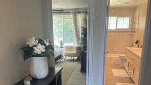 A hallway leading to a bedroom with a bed, a chair, and a potted plant. There is a bathroom with a bathtub, toilet, and sink visible through an open door. - Charming 4-br Wine Country Retreat — Walk Downtown Solvang + BBQ & Fireplace (Solvang)