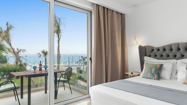 A hotel room with a large sliding glass door leading to a balcony with a view of the sea. There is a bed with a tufted headboard, a small table, and a chair on the balcony. - Le Palme Beach Rhodes- Pool Heart Villa (Χαράκι)