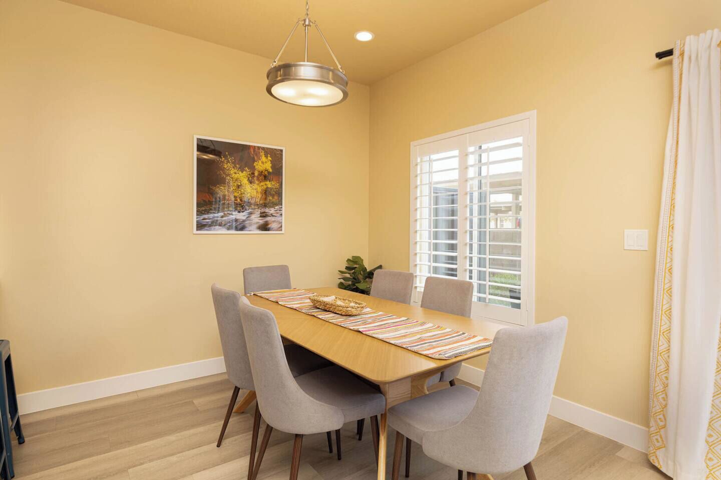 A dining area with a wooden table, grey chairs, a hanging light fixture, a window with blinds, and a framed picture on the wall.