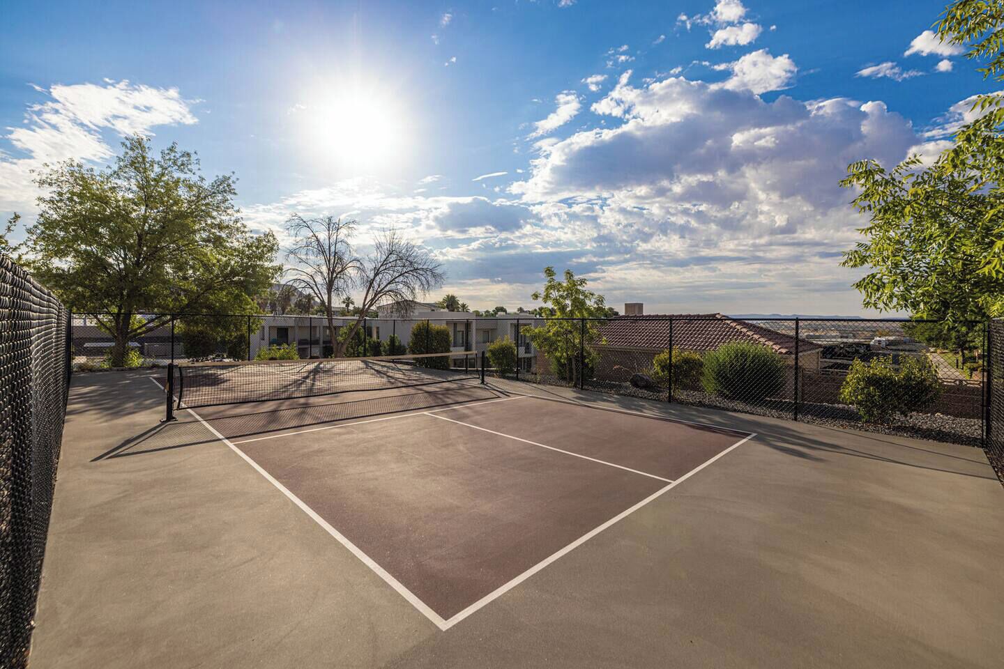 An outdoor tennis court with a chain-link fence, surrounded by trees and buildings.
