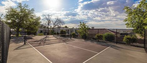 An outdoor tennis court with a chain-link fence, surrounded by trees and buildings.