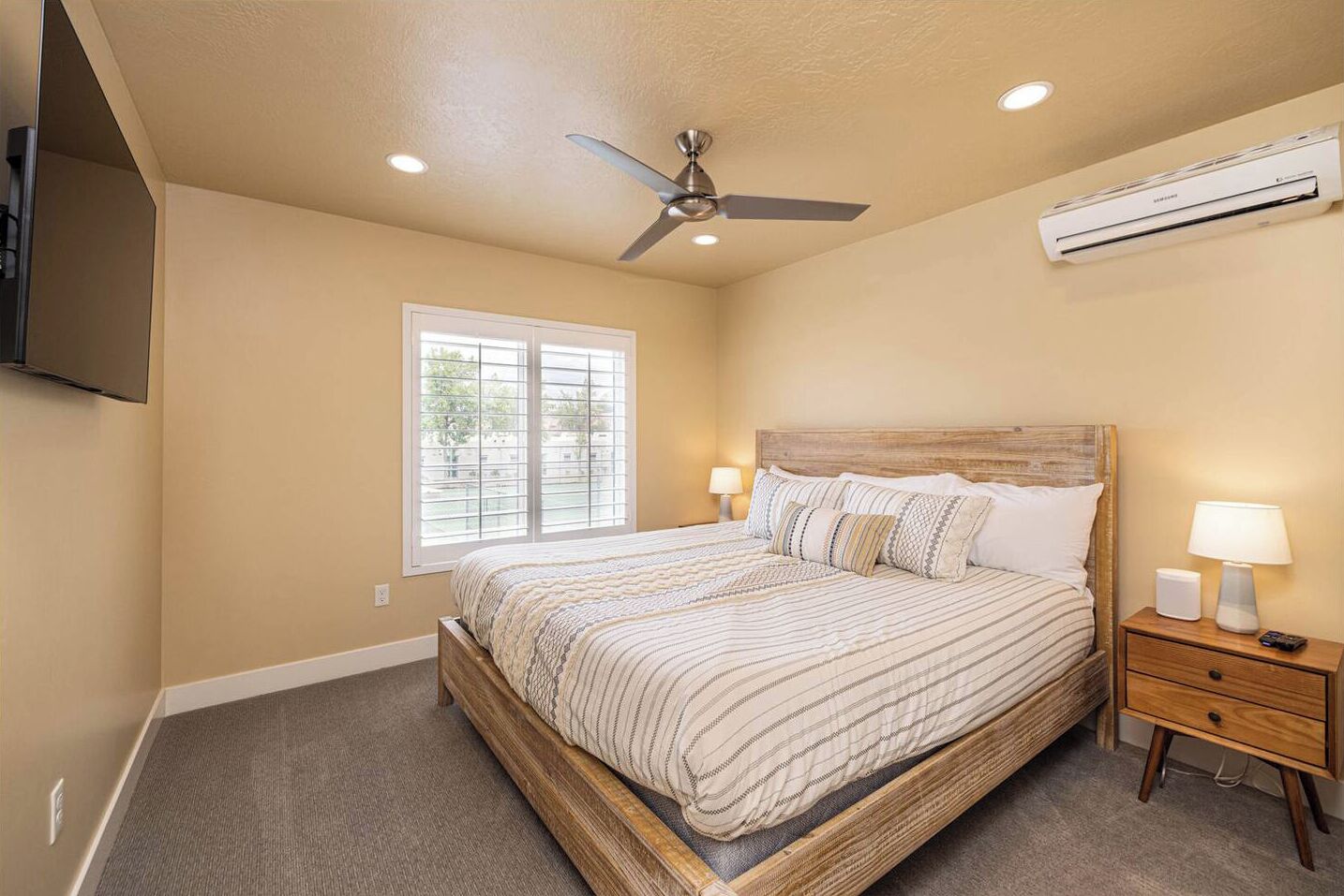 A bedroom with a bed, a ceiling fan, an air conditioning unit, a window, and a nightstand with a lamp.