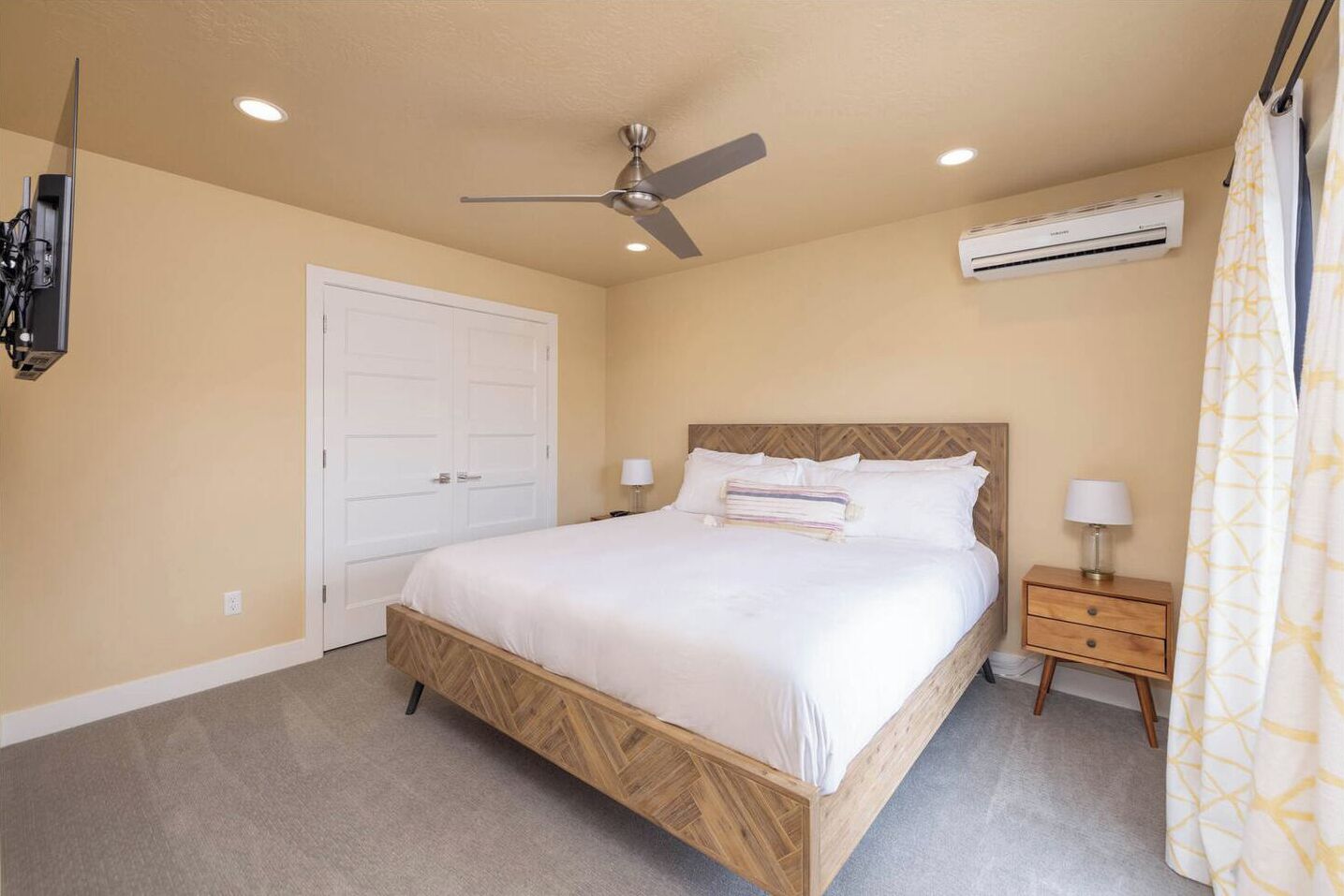 A bedroom with a bed, a ceiling fan, an air conditioning unit, a bedside table with a lamp, and a door.
