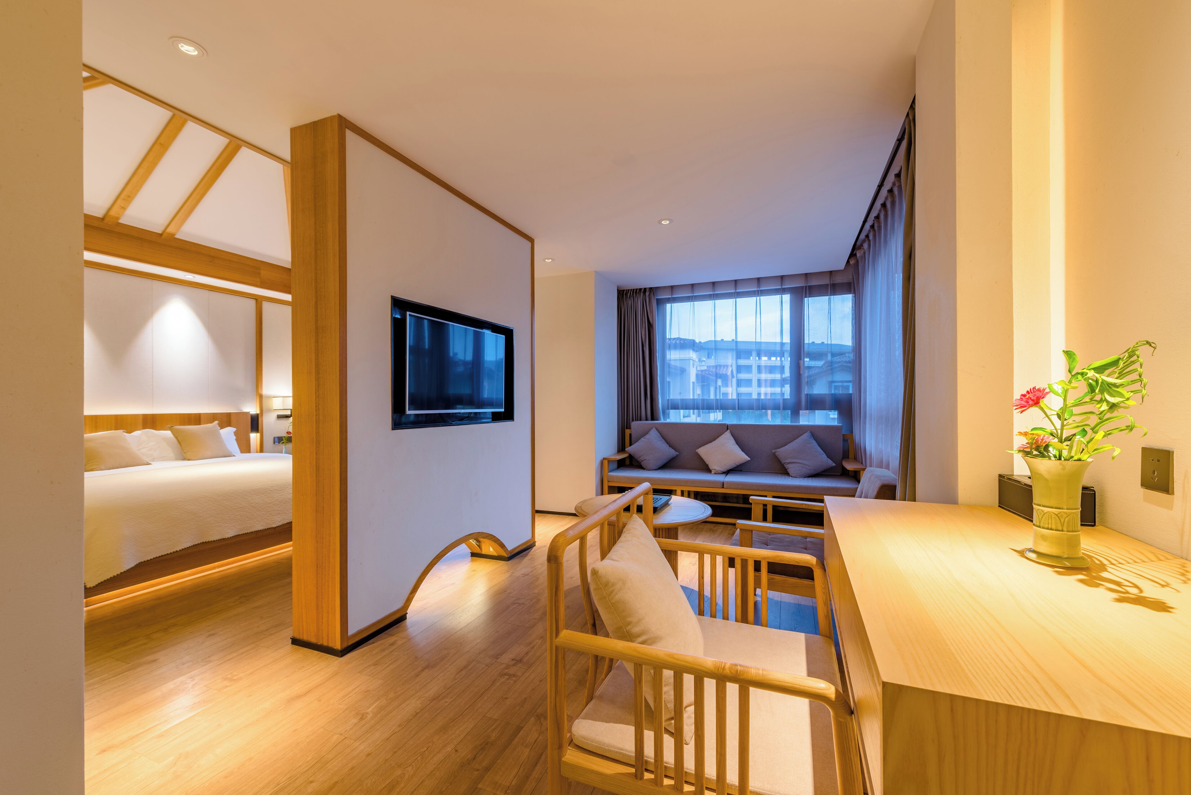 Habitación de hotel moderna con piso de madera, cama con ropa blanca, sofá con cojines, mesa de comedor y televisor montado en la pared.