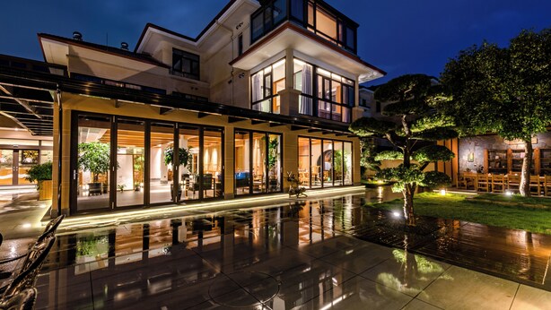Une maison moderne à deux étages, dotée de grandes fenêtres et agrémentée d’un bassin dont la surface réfléchit la lumière, situé à l’avant.
