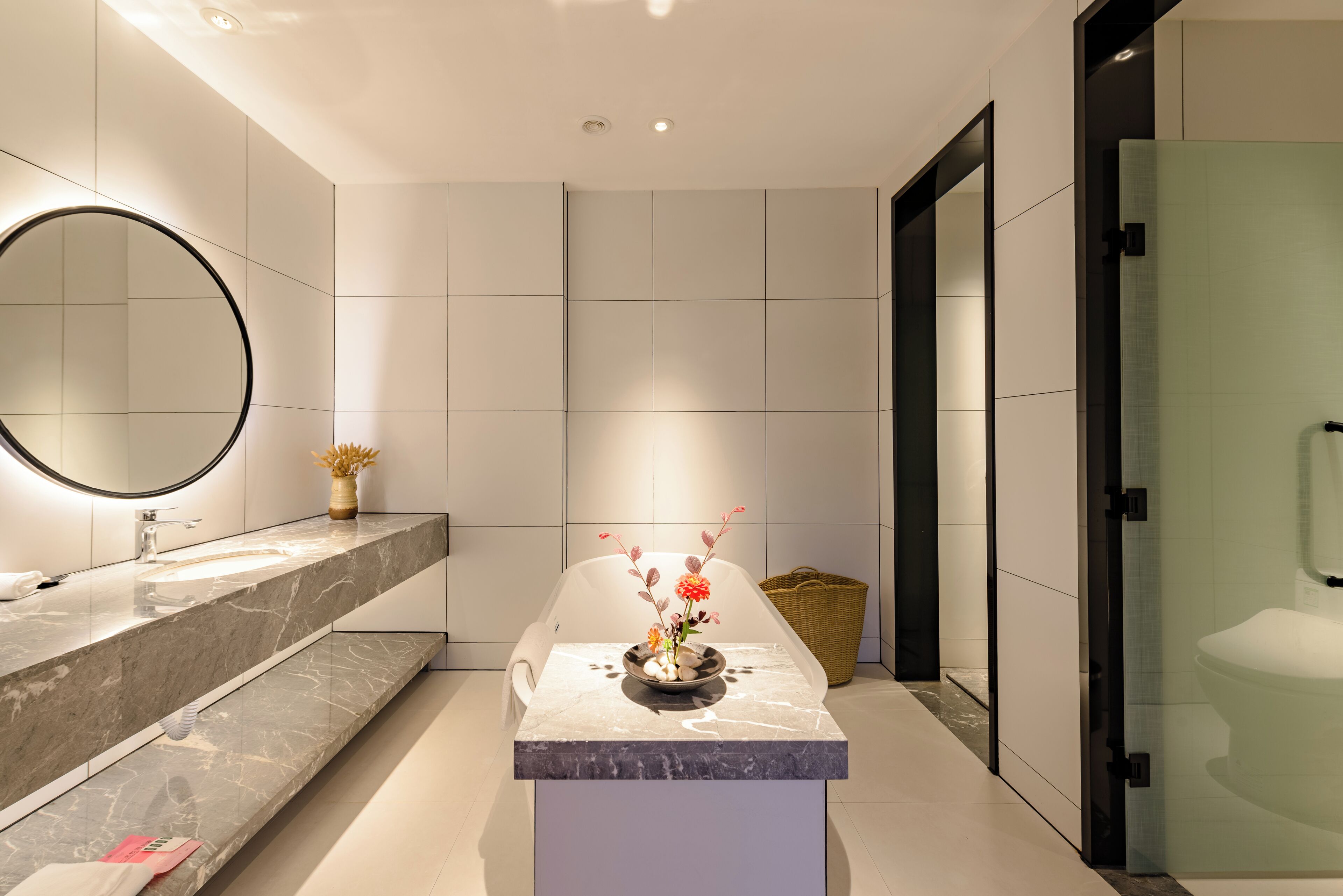 A modern bathroom with a marble vanity, a round mirror, and a vase with flowers.
