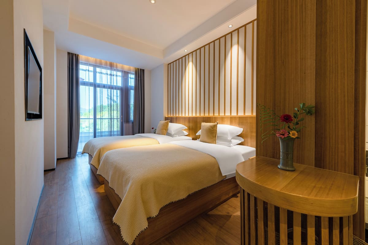 A hotel room with a large bed, a wooden headboard, a bedside table with a vase of flowers, and a view of the outdoors through the window.