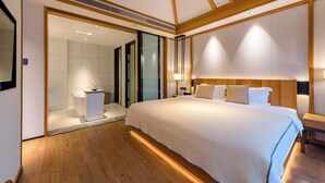 Une chambre d’hôtel moderne avec un grand lit, des poutres apparentes en bois au plafond, une petite table de chevet et une salle de bain visible à travers une porte ouverte.