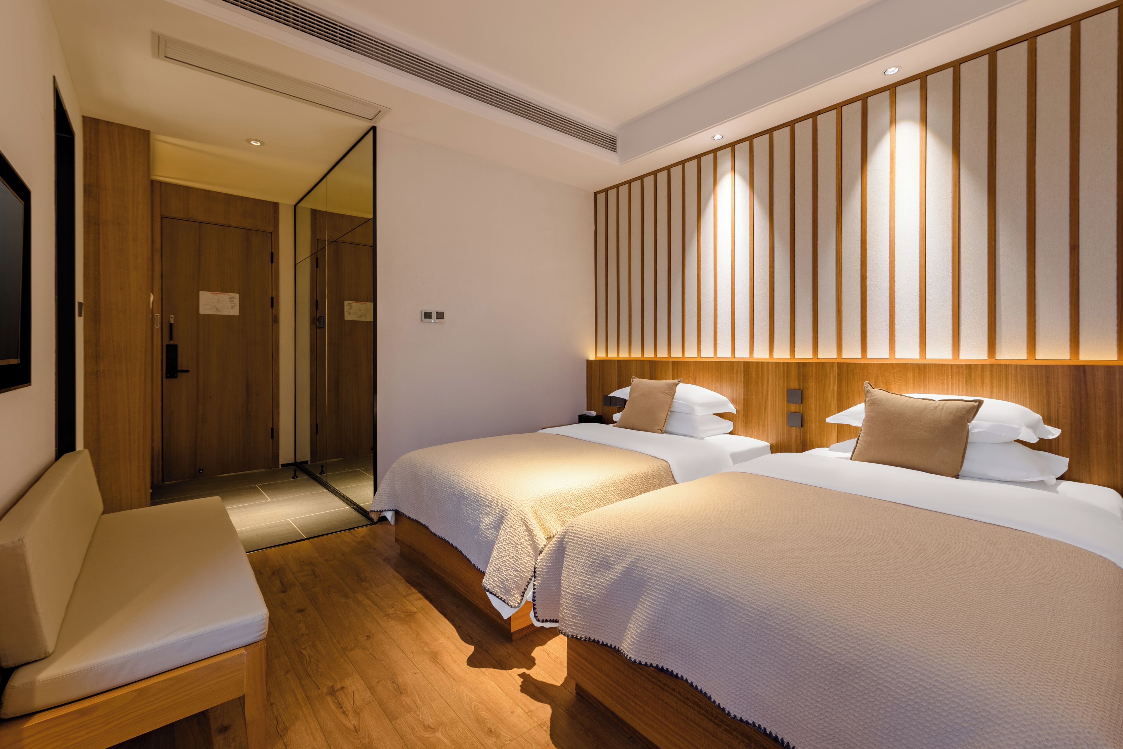 A hotel room with two beds, a wooden bench, a large mirror, and a wooden panel wall.