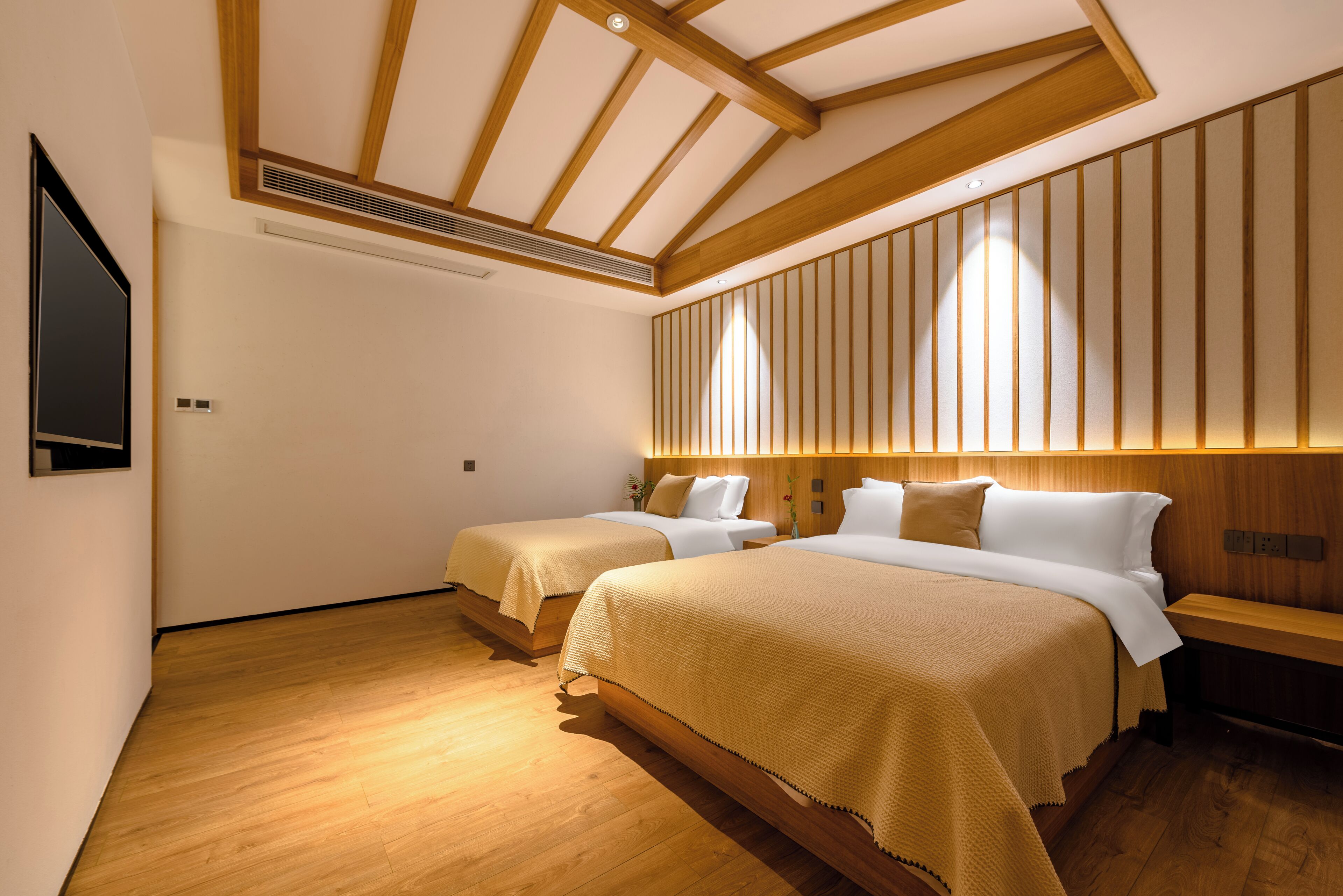 Habitación de hotel con dos camas, suelo de madera, televisor de pantalla plana montado en la pared y cabecero de madera con listones verticales.