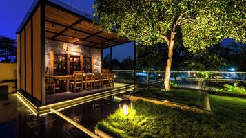Un espacio moderno para comer al aire libre con pared de piedra, muebles de madera y un árbol.