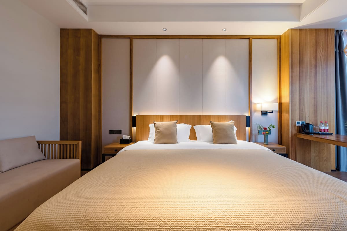 A hotel room with a large bed, a wooden headboard, a bedside table with a lamp, and a small nightstand.