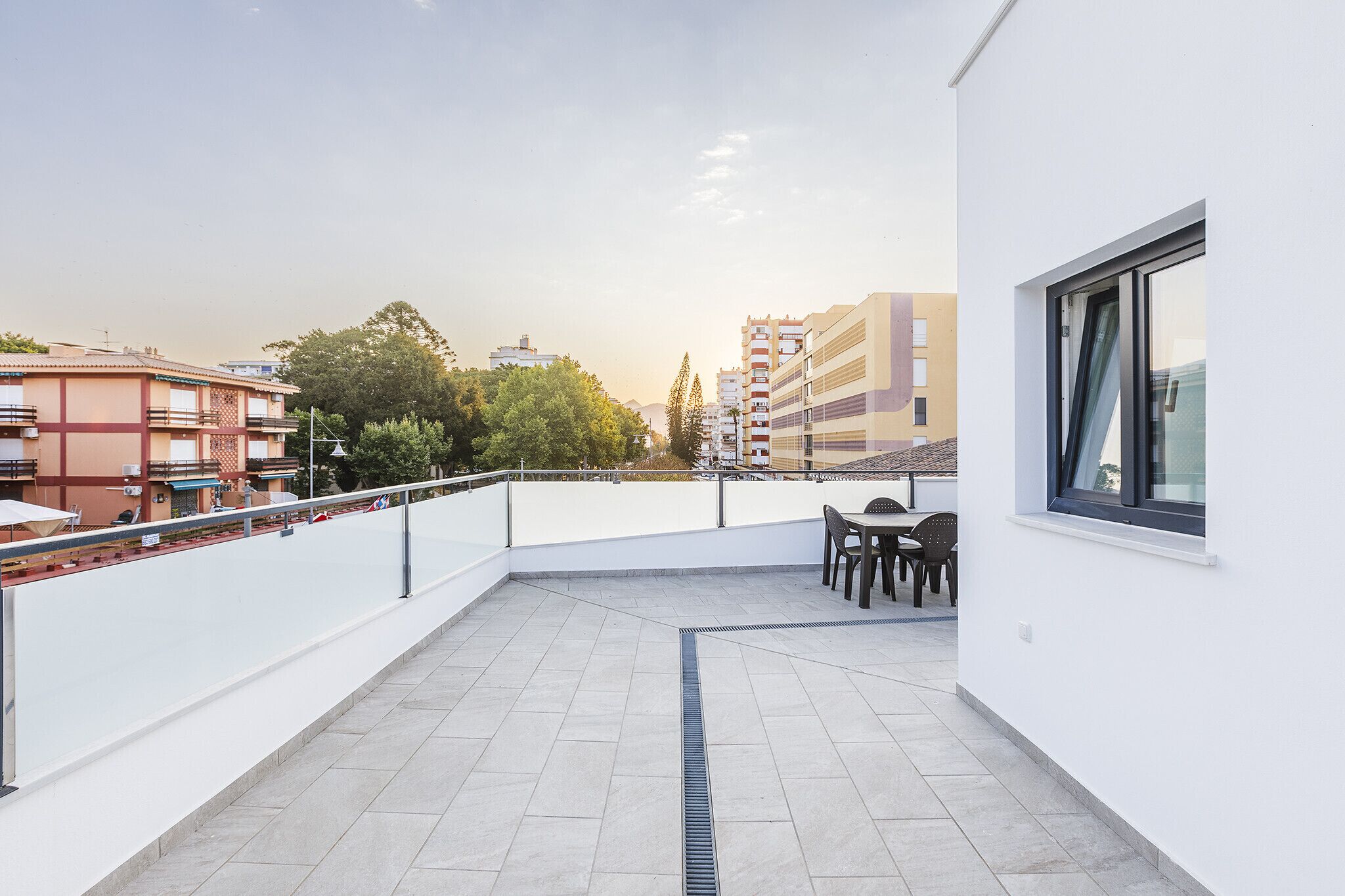 A rooftop terrace with a table and chairs, a view of surrounding buildings, and a clear sky.