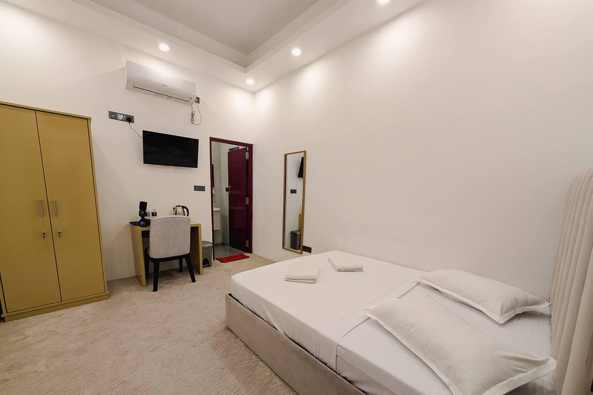 A hotel room with a bed, a desk with a chair, a wardrobe, a TV mounted on the wall, and a mirror.