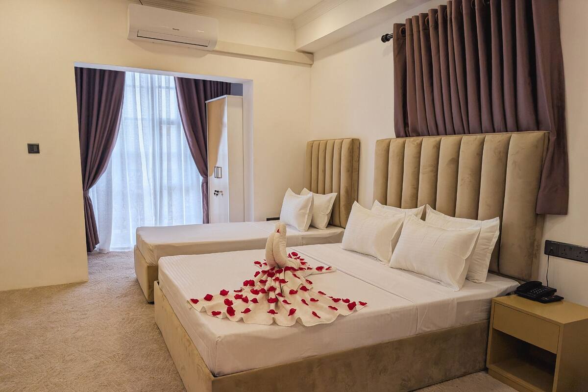 A hotel room with two beds, a large window with curtains, an air conditioning unit, and a bedside table with a phone.