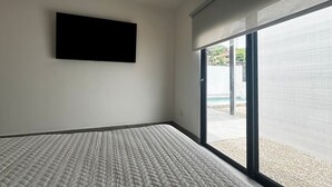 A bedroom with a bed, a flat-screen TV, and a large sliding glass door leading to an outdoor area. - Ocean Villas Modern Home w/ Pool (Coco)