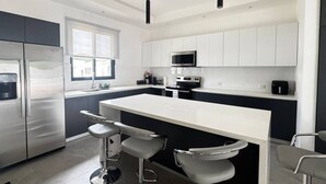 A modern kitchen with a central island, bar stools, stainless steel appliances, and white cabinetry. - Ocean Villas Modern Home w/ Pool (Coco)