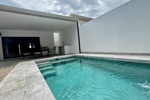 An outdoor swimming pool with a tiled deck, a seating area with chairs, and a small kitchen area with a sink and counter.
