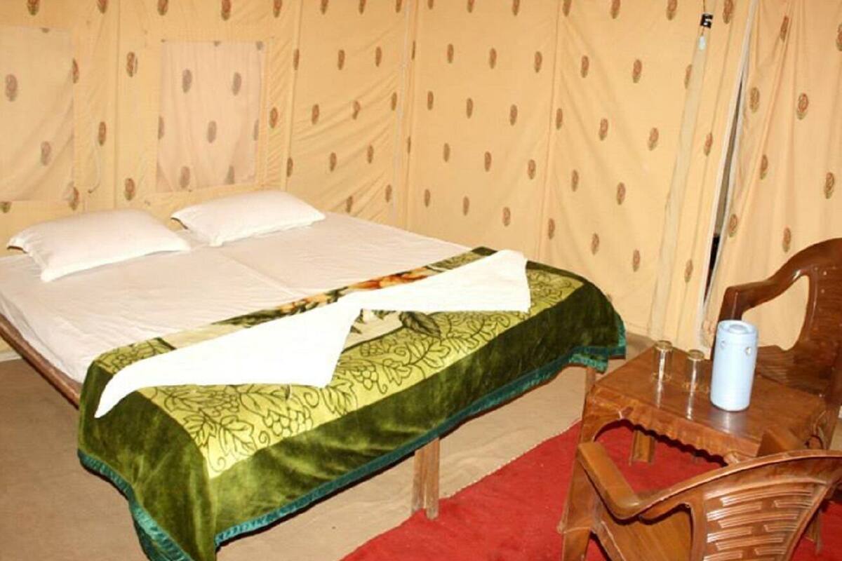 A single bed with a green and gold bedspread, a wooden chair, and a small table with a blue object on it.
