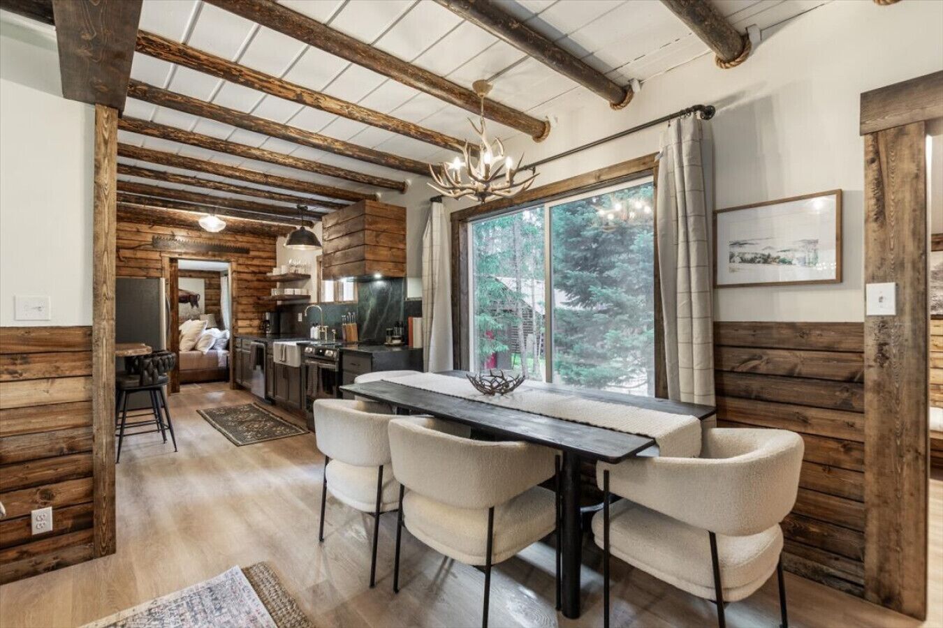 A rustic interior with exposed wooden beams, a dining area with a table and chairs, a kitchen with modern appliances, and a large window offering a view of trees.