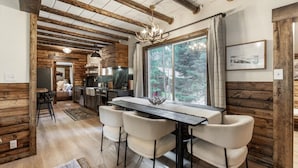 A rustic interior with exposed wooden beams, a dining area with a table and chairs, a kitchen with modern appliances, and a large window offering a view of trees.