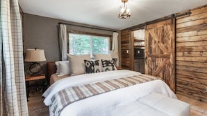 A bedroom with a large bed, a wooden wardrobe, a nightstand with a lamp, and a window with curtains.