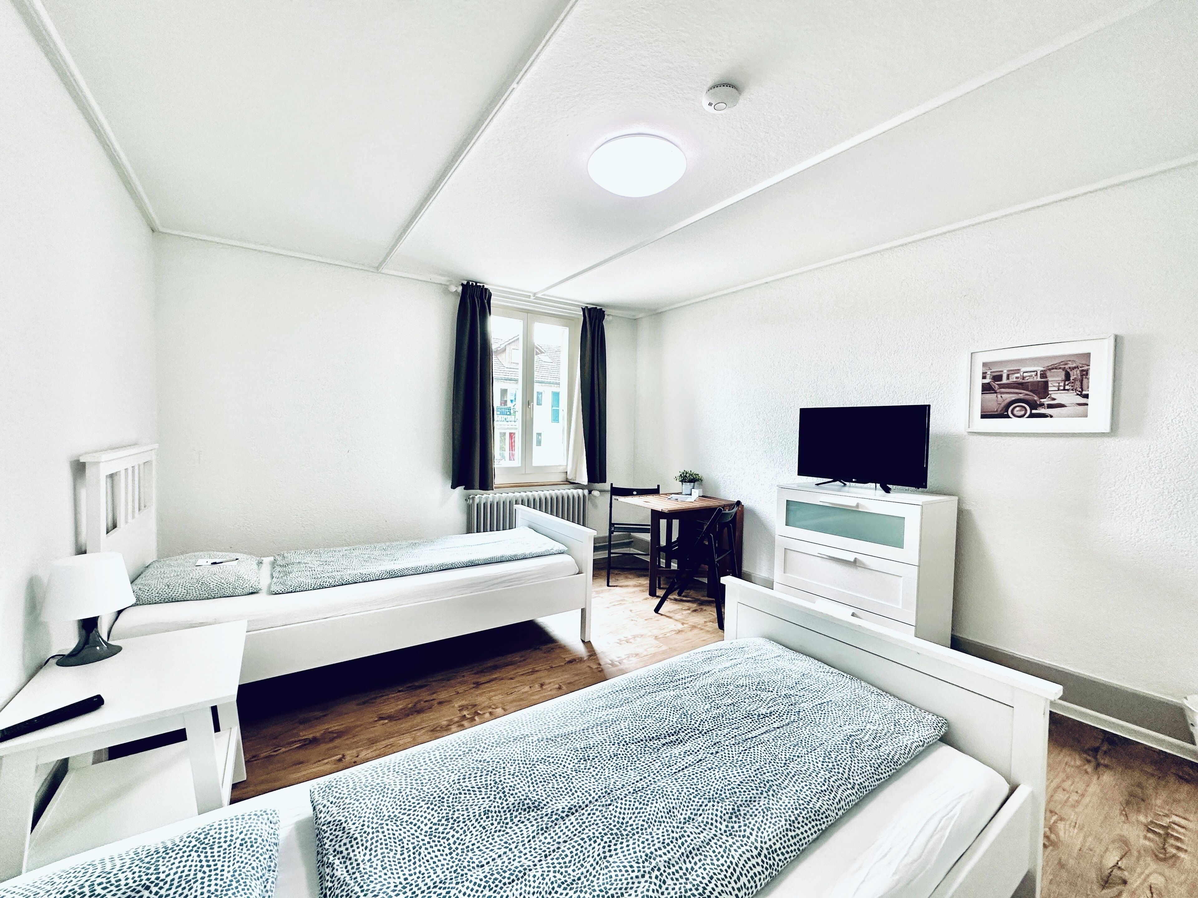 Una habitación pequeña y limpia con dos camas, un escritorio, una silla, un televisor y una ventana con cortinas.