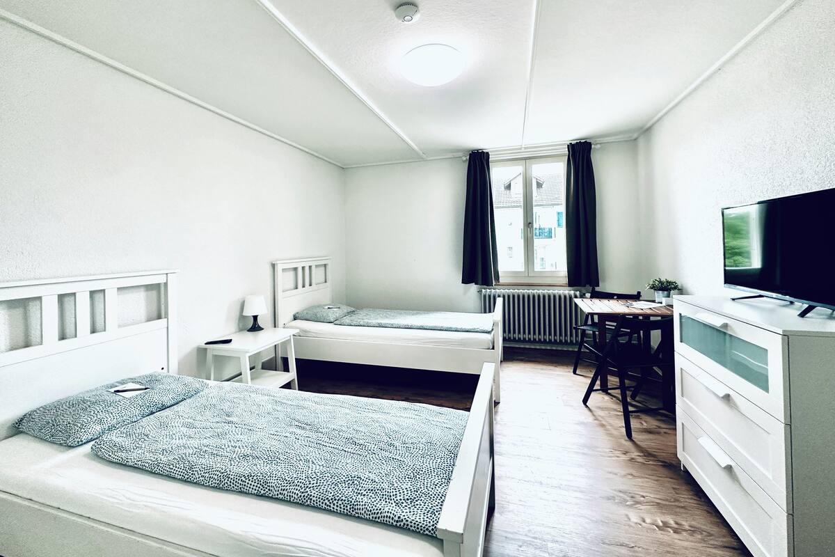 Una habitación pequeña con dos camas individuales, un escritorio, una silla, un radiador y un televisor.