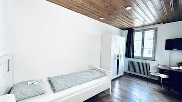 Una habitación moderna con techo de madera, sofá blanco con cojines estampados, armario blanco, radiador y ventana con cortinas.