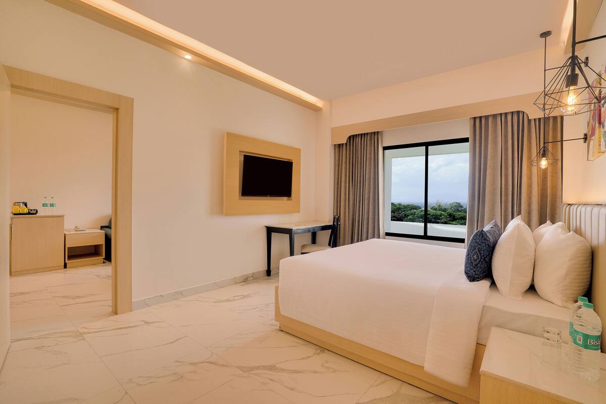 A hotel room with a large bed, a TV mounted on the wall, a desk, and a window with curtains.