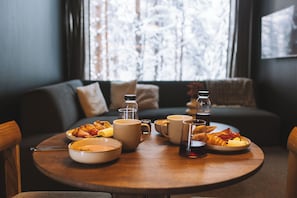 Une table à manger avec des aliments pour le petit-déjeuner, un canapé et une fenêtre donnant sur des arbres.