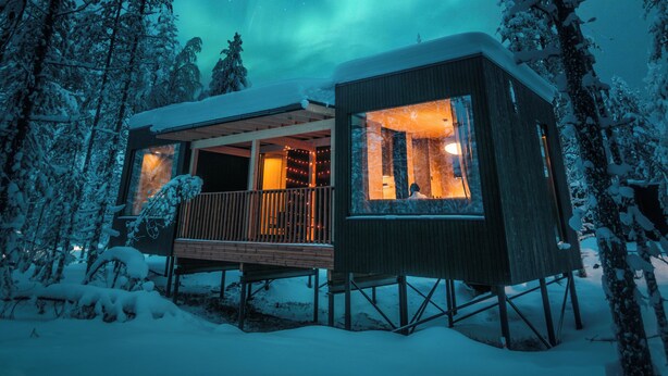 Eine Holzhütte mit einer Holzterrasse, umgeben von schneebedeckten Bäumen, unter einem Nachthimmel mit Sternen und Polarlichtern.