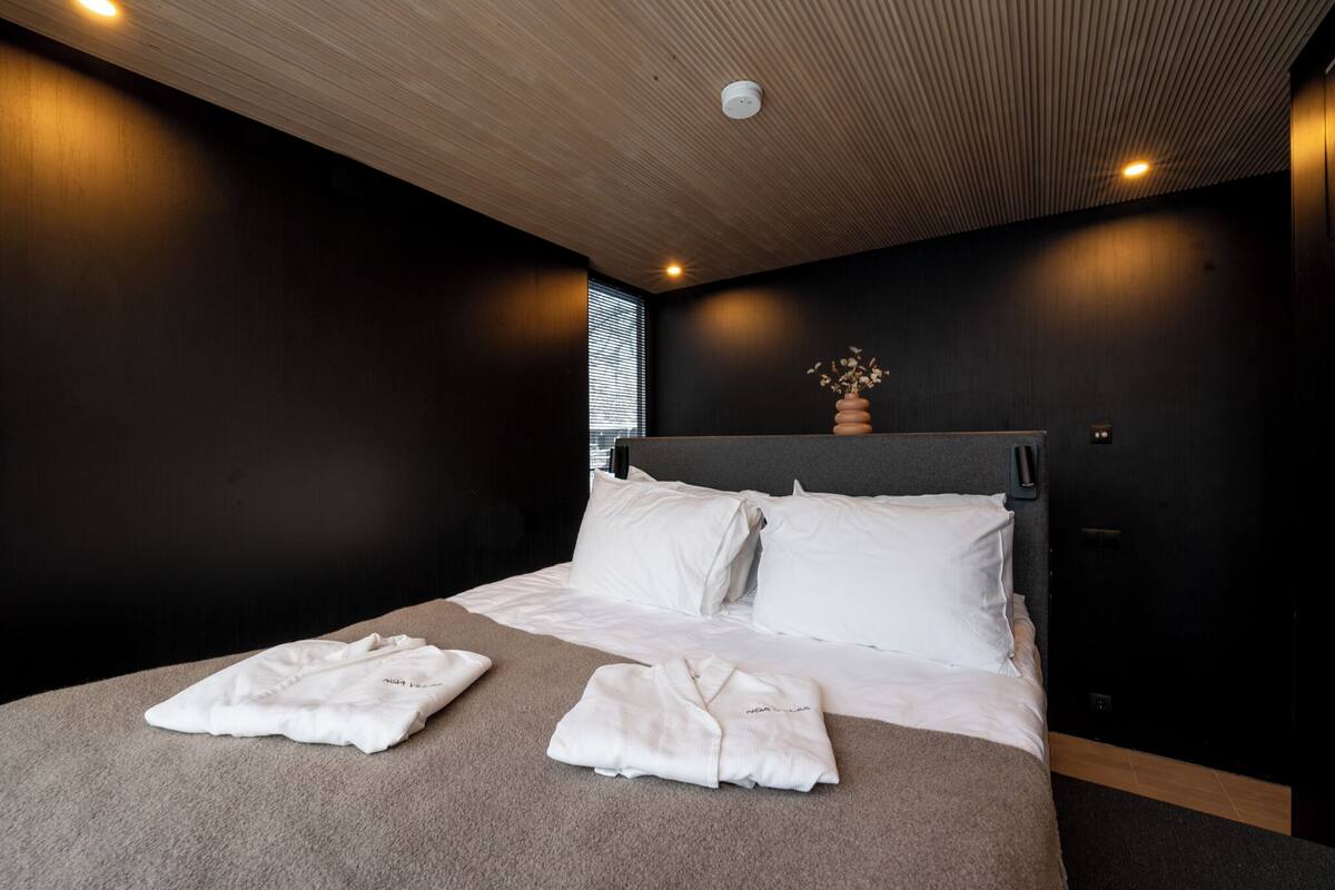 Habitación de hotel moderna con una cama grande, ropa de cama blanca, techo de madera y un pequeño jarrón en la cabecera.