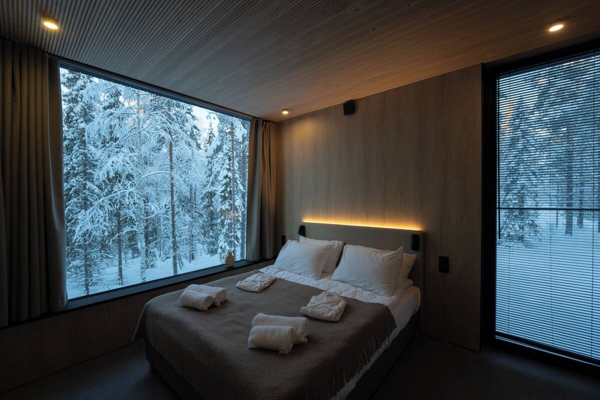 Habitación de hotel moderna con un ventanal grande que ofrece vistas a un bosque nevado, una cama bien hecha y una cálida iluminación ambiental.
