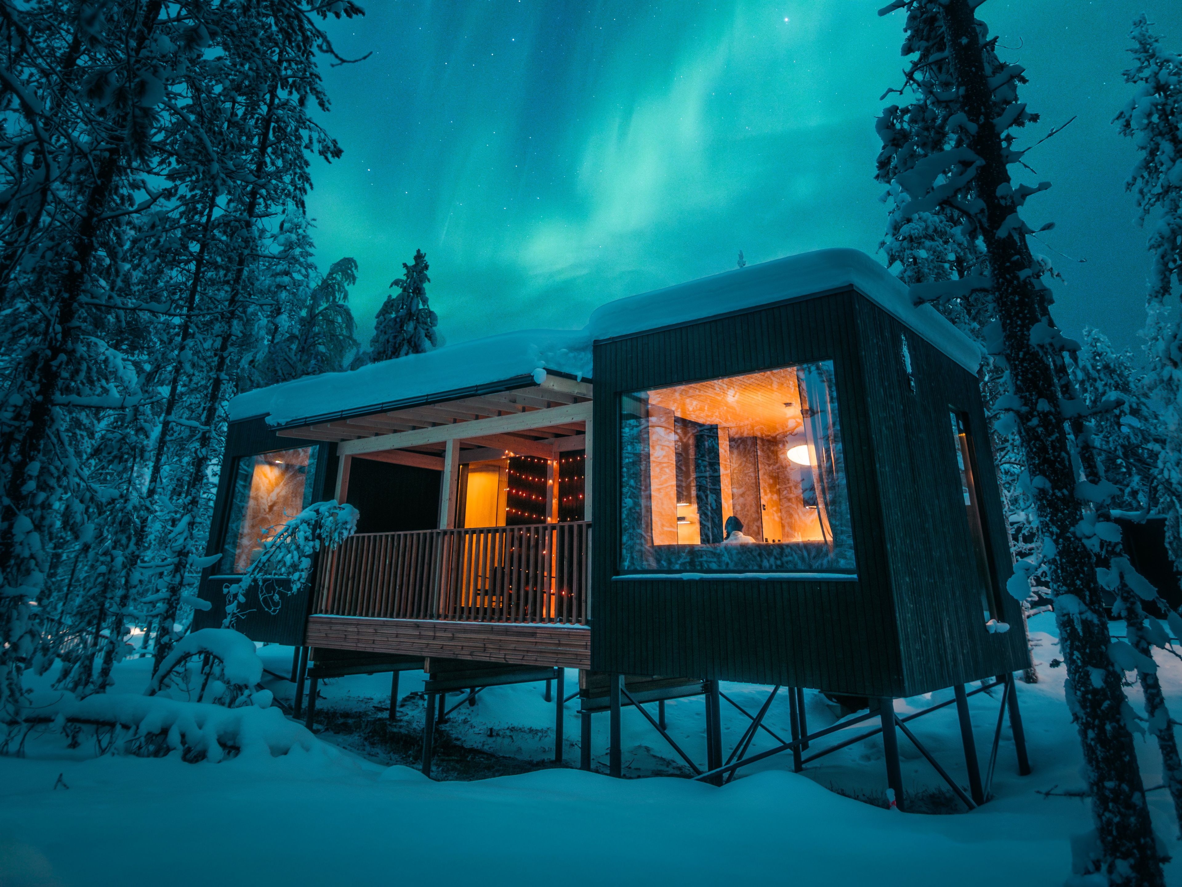 Una baita con un balcone, circondata da alberi innevati, sotto un cielo notturno con aurore boreali visibili.