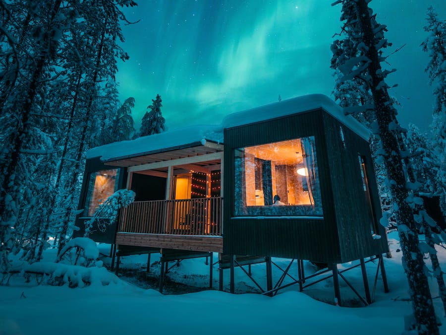 A cabin with a balcony, surrounded by snow-covered trees, under a night sky with visible aurora borealis.