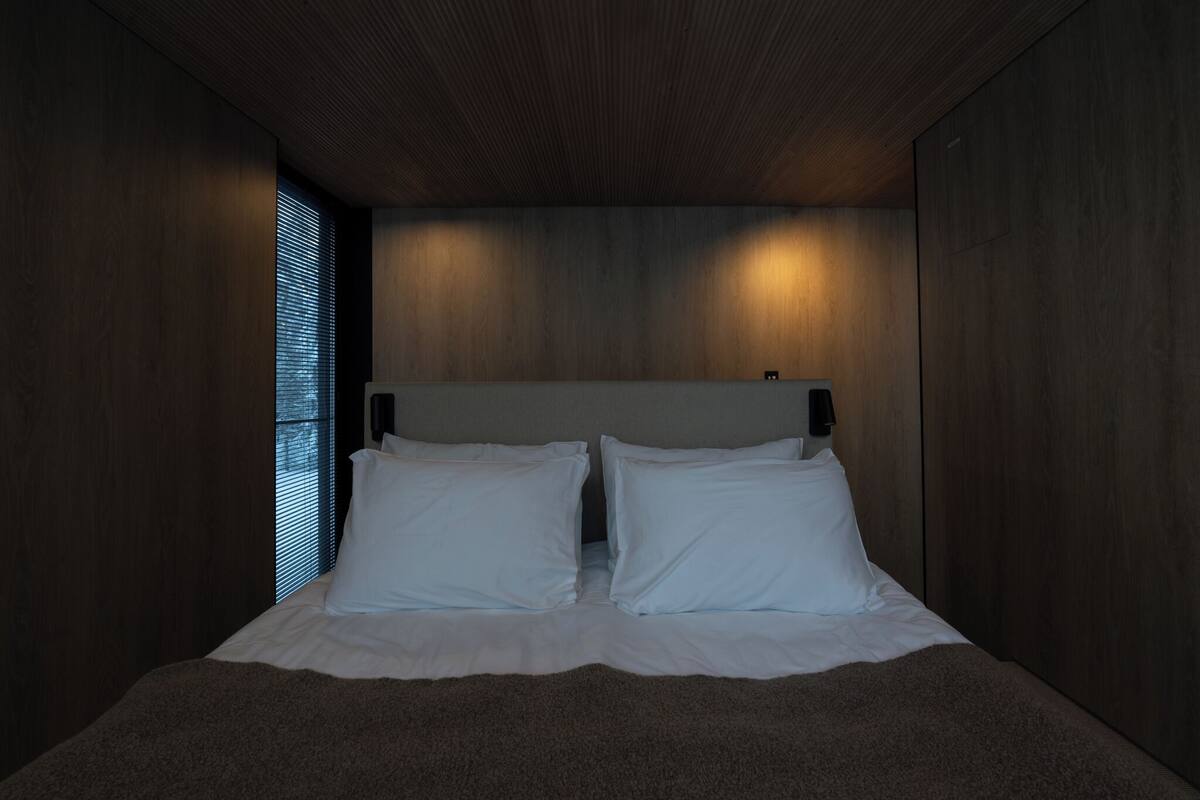 Un dormitorio moderno con cabecera de madera, dos almohadas blancas y una ventana con persianas.