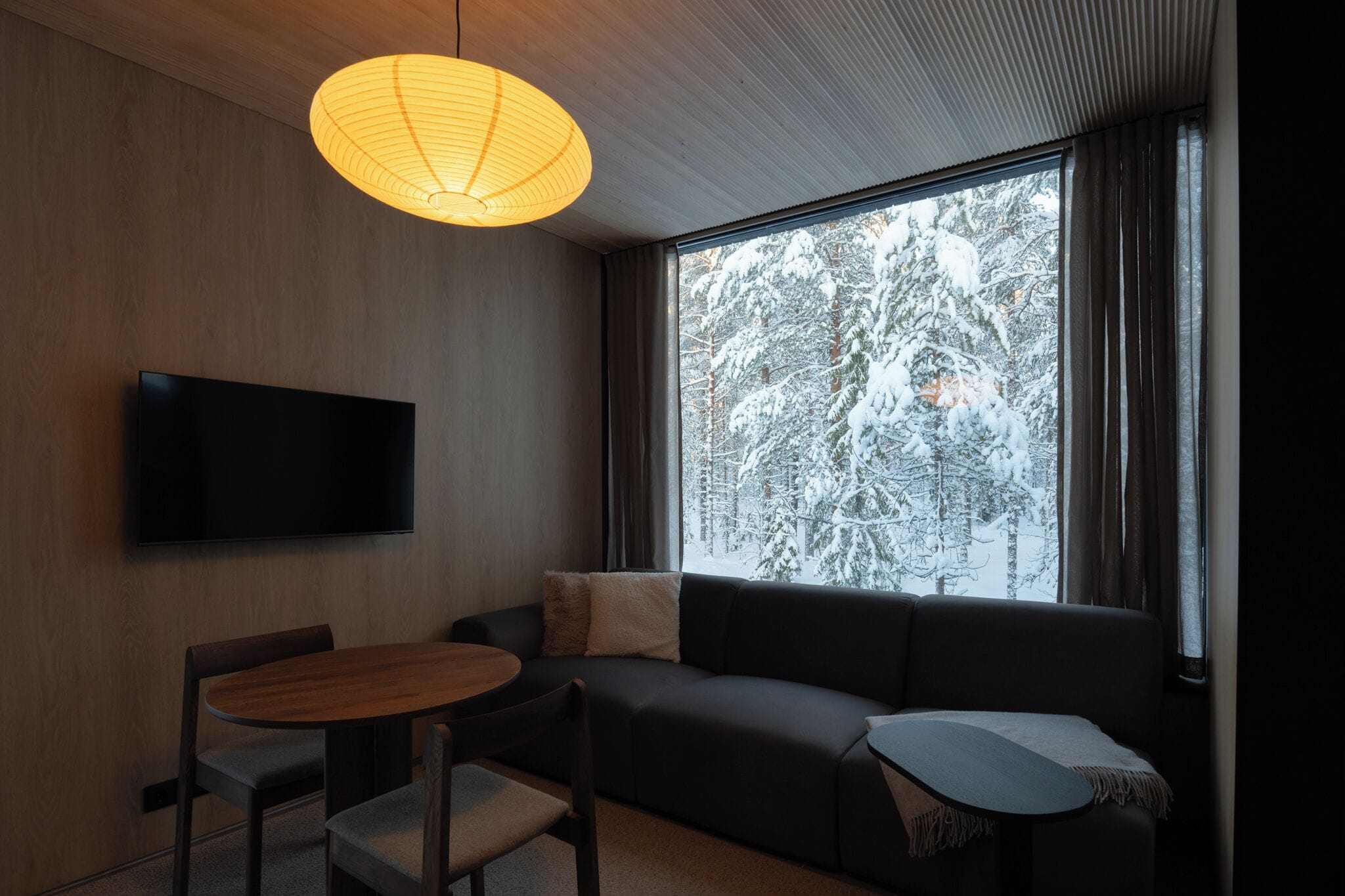 Uma sala de estar moderna com um sofá, uma mesa de madeira redonda e uma TV fixada na parede. Uma janela grande oferece vista para árvores cobertas de neve.