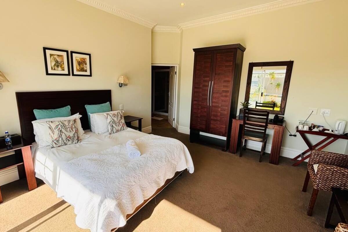 A hotel room with a large bed, a wooden wardrobe, a desk with a chair, and a small table with a lamp.