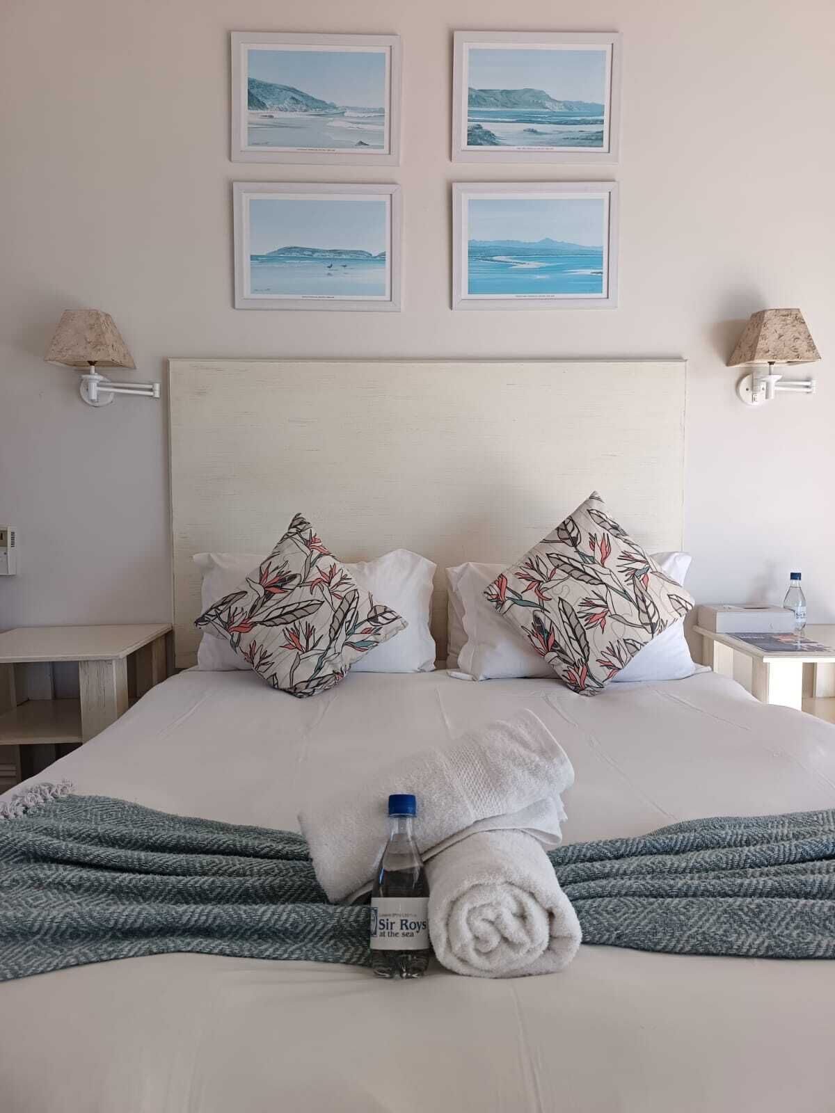 Un lit bien fait, agrémenté d’oreillers à motifs floraux, d’une serviette roulée et d’une bouteille de Sir Roy, avec vue sur la mer. Au-dessus du lit, on peut voir des photos encadrées représentant des paysages côtiers.
