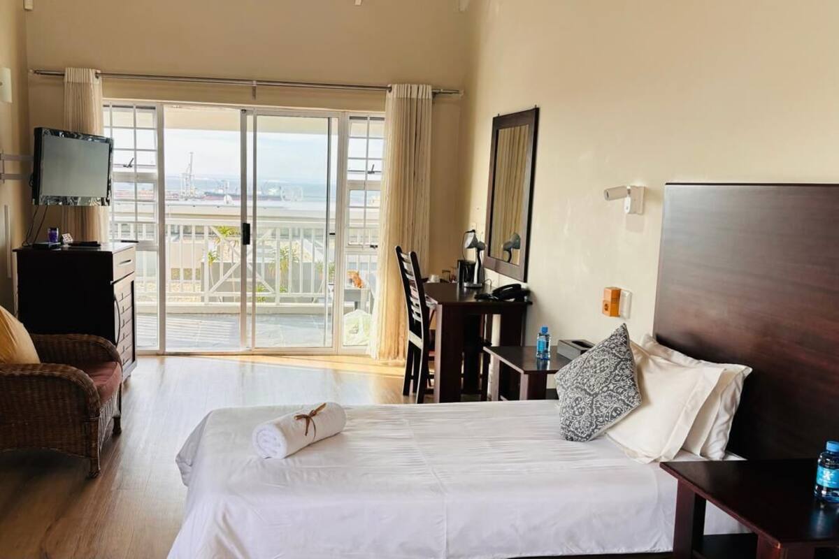 A hotel room with a bed, a chair, a desk, a TV, and a balcony with a view of the sea.