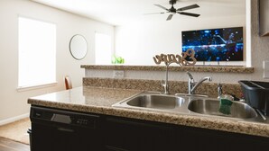 Una cocina con encimeras de granito, lavamanos de acero inoxidable, lavavajillas, ventilador de techo y televisor montado en la pared.