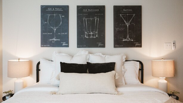 Una cama bien hecha con almohadillas blancas y negras, flanqueada por dos lámparas de noche. Sobre la cama, tres dibujos enmarcados de copas de vino titulados 'Gin & Tonic', 'Old Fashioned' y 'Martini'.
