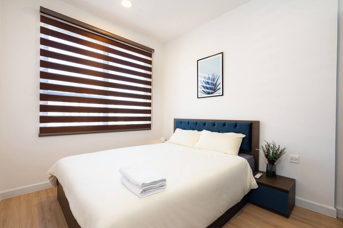 A neatly made bed with white linens and a folded towel, a blue upholstered headboard, a bedside table with a vase of flowers, and a large window with horizontal blinds.