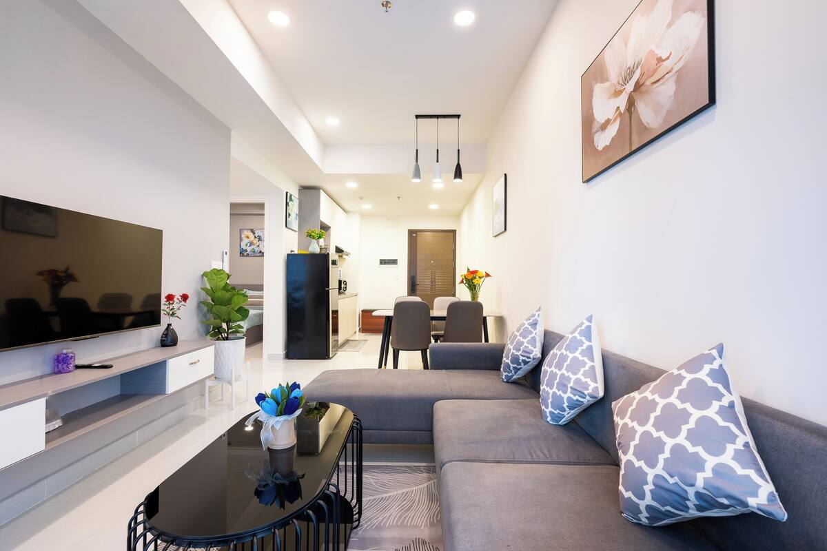 A modern living room with a flat-screen TV, a grey sofa, and a dining area in the background.