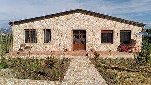 A stone building with a central door, two windows on each side, and a covered outdoor seating area with red chairs.