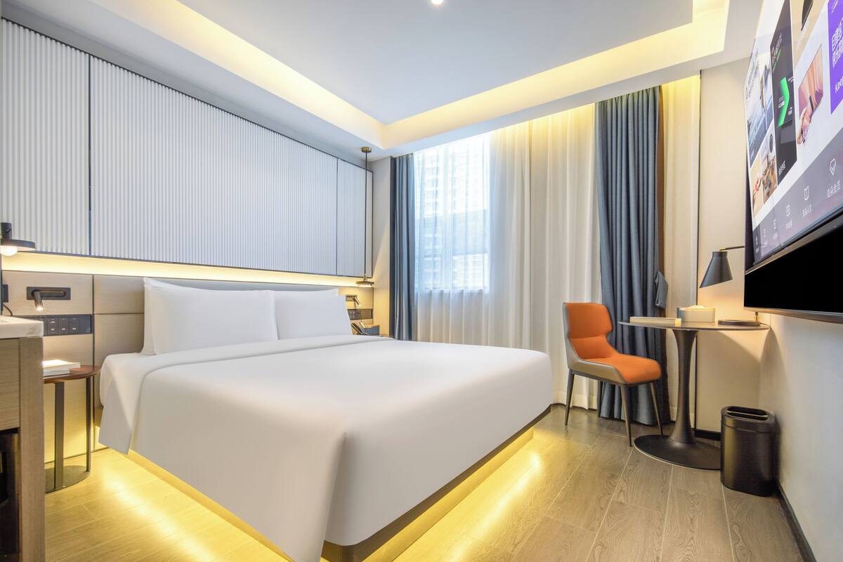 Una habitación de hotel moderna con una cama grande, un escritorio, una silla, un televisor y una ventana con cortinas.