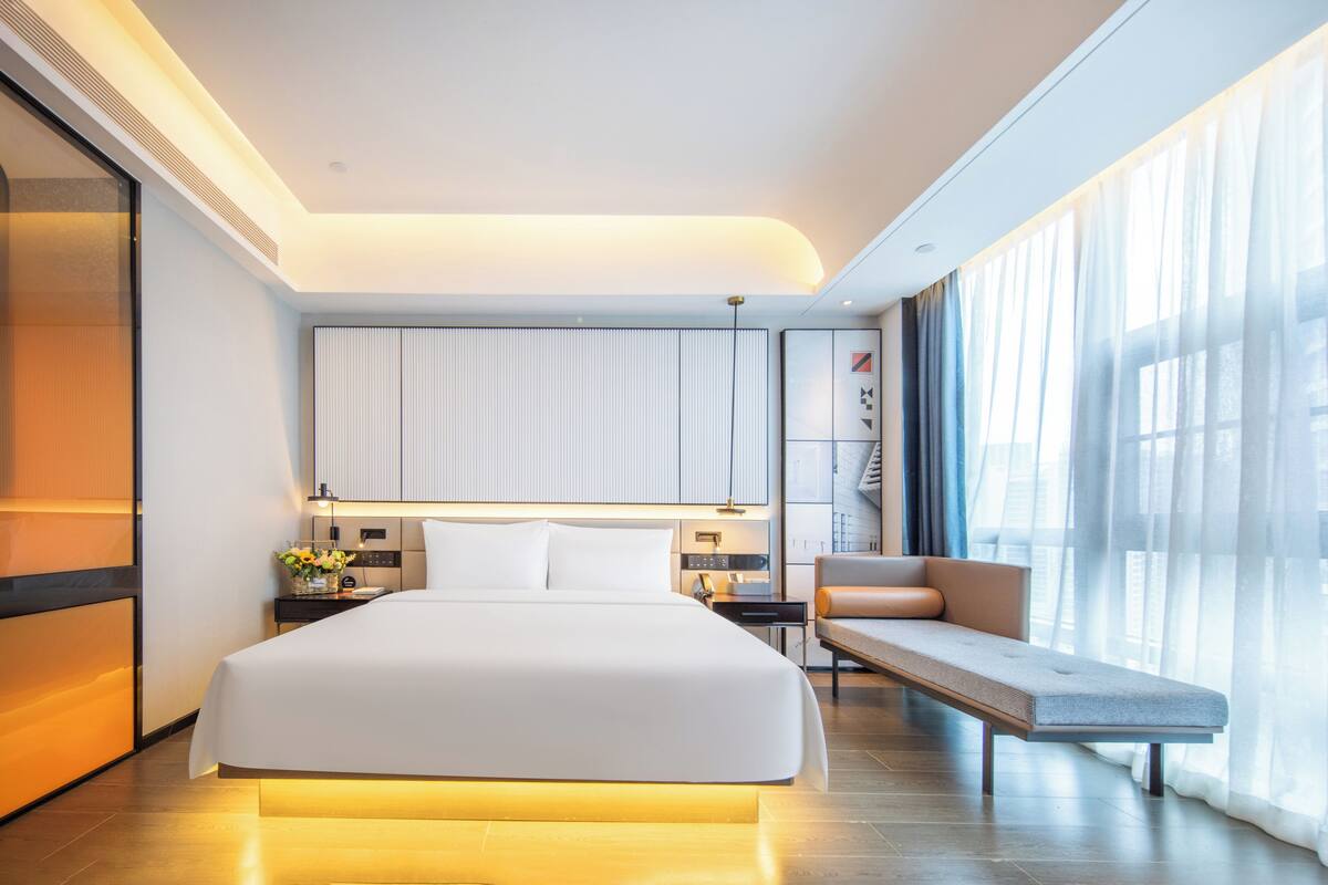 Una habitación de hotel moderna con una cama grande, mesitas de noche, un banco y vistas a la ciudad a través de amplios ventanales.