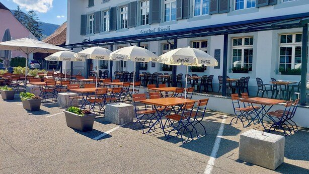 An outdoor café with tables and chairs, umbrellas, and a building with a sign reading "Gasthof Rössli". - ZIMMERzuVERMIETEN.ch Oensingen (Oensingen)
