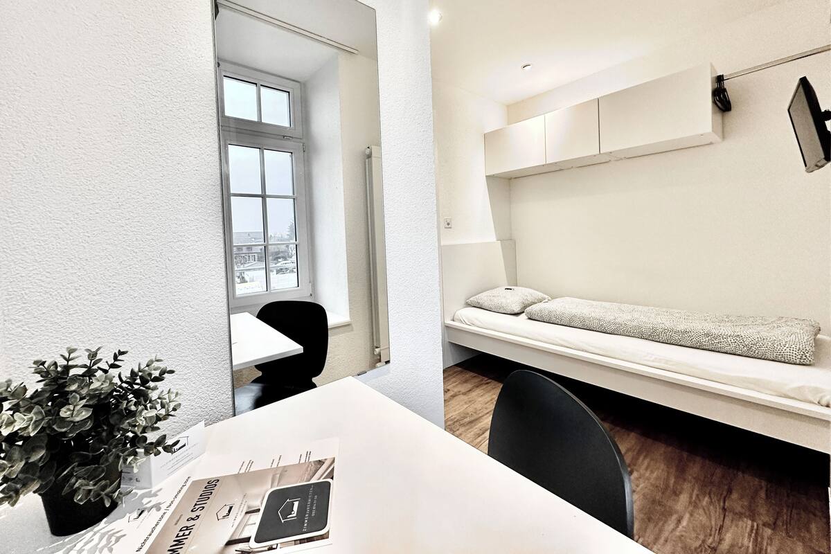 Une chambre moderne avec un lit blanc, une petite table ornée d’une plante et une fenêtre.